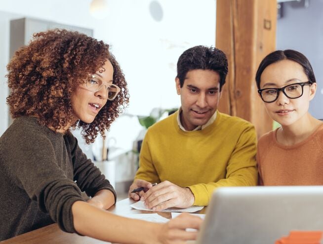 a curly haired woman wearing glasses presenting something to her work colleagues, all sat side by side at a desk in an office