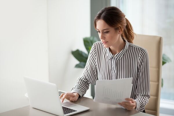 a woman wearing a striped shirt sat a desk in a bright room looking down at her open laptop, pressing a key whilst holding a piece of paper