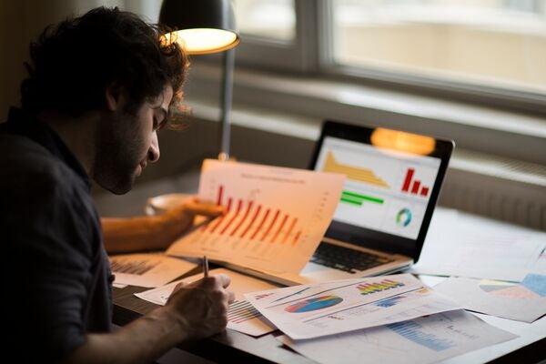 a man sat at a desk holding a piece of paper with charts on whilst having his laptop open showing financial reports