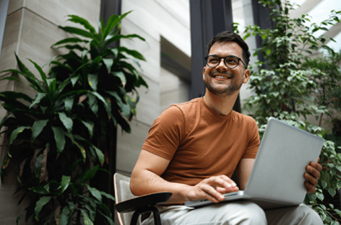 a smiling man wearing glasses and an orange t-shirt, looking into the distance whilst sat down in an office with his laptop open on his lap surrounded by green house plants