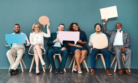 6 people sat down in a row smiling and holding thought bubbles made of cardboard