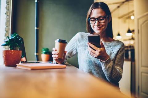 a woman sat at a table holding a takeaway cup of coffee whilst looking down and smiling at mobile phone